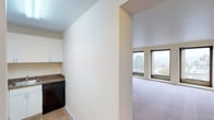 This single-floor residence offers a clean, functional layout designed for comfortable urban living. The kitchen is efficiently arranged with white cabinetry, dark granite-style countertops, hardwood-style flooring, and a full suite of black appliances including a refrigerator, electric range, microwave, and dishwasher. It opens into a hallway that leads to the expansive living area, where plush carpeting and warm, neutral-toned walls create a welcoming atmosphere. A wall of large picture windows fills the living space with natural light and frames impressive elevated views of the surrounding cityscape and hills beyond. The bedroom continues the carpeted, light-toned aesthetic and is equipped with a ceiling fan, two large windows offering the same scenic city views, and a generous reach-in closet with a full-width hanging rod and bifold doors. The bathroom features a classic white-tiled tub and shower combination with chrome fixtures, set alongside a vanity with wood-toned cabinetry. Throughout the unit, the consistent use of soft cream and beige tones, along with ample natural light, gives the space a bright, airy, and cohesive feel suited for a variety of lifestyles.