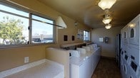 This single-floor laundry facility is designed for efficient, communal use, offering a clean and functional environment for residents or guests. The room features a continuous row of top-load washing machines neatly aligned along one wall, providing generous washing capacity. A large built-in utility sink with a speckled countertop sits prominently near the entrance, ideal for hand-washing or pre-treating garments. An extended folding counter runs alongside the machines, offering convenient space for sorting and organizing laundry. Natural light floods the room through multiple windows, giving the space a bright and airy feel with views of the surrounding area. The warm, neutral-toned walls and dark tile flooring create a practical yet welcoming atmosphere. Overhead, a ceiling fan with a light fixture and an additional globe pendant light ensure the space is well-lit at all times. Informational notices and a clock are mounted on the walls, keeping users informed and on schedule. The overall layout is thoughtfully organized to maximize workflow and ease of use, making this laundry facility a highly functional shared amenity.