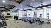 This single-floor fitness center offers a comprehensive workout environment designed for both cardio and strength training. The space is distinguished by its elegant coffered ceiling with dark-trimmed box detailing and recessed lighting, lending a refined aesthetic uncommon in typical gym settings. A ceiling fan adds to the comfort of the room.

Natural light filters in through multiple windows dressed with white plantation shutters, brightening the space and creating an open, airy atmosphere. Patterned carpet tiles in neutral tones cover the floor throughout, while rubber mats are positioned beneath the cardio equipment for added stability and protection.

The cardio section is well-appointed with a row of treadmills, elliptical machines, and a stationary bike, each equipped with individual display monitors. A dedicated free weights area features a multi-tiered dumbbell rack alongside a weight bench and cable machine, catering to a range of strength training needs. Large wall-mounted mirrors in the free weights section enhance the sense of space and allow users to monitor their form. The overall ambiance is polished and functional, offering a welcoming setting for a variety of fitness routines.
