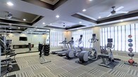This single-floor fitness center offers a comprehensive workout environment designed for both cardio and strength training. The space is distinguished by its elegant coffered ceiling with dark-trimmed box detailing and recessed lighting, lending a refined aesthetic uncommon in typical gym settings. A ceiling fan adds to the comfort of the room.

Natural light filters in through multiple windows dressed with white plantation shutters, brightening the space and creating an open, airy atmosphere. Patterned carpet tiles in neutral tones cover the floor throughout, while rubber mats are positioned beneath the cardio equipment for added stability and protection.

The cardio section is well-appointed with a row of treadmills, elliptical machines, and a stationary bike, each equipped with individual display monitors. A dedicated free weights area features a multi-tiered dumbbell rack alongside a weight bench and cable machine, catering to a range of strength training needs. Large wall-mounted mirrors in the free weights section enhance the sense of space and allow users to monitor their form. The overall ambiance is polished and functional, offering a welcoming setting for a variety of fitness routines.