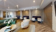 Located at 450 Perimeter Dr in Midlothian, Virginia, this thoughtfully designed single-floor amenity space offers a sophisticated and inviting environment for socializing and relaxation. The layout flows seamlessly between a well-appointed kitchen and bar area and a spacious lounge, creating distinct zones for gathering and unwinding.

The kitchen and bar area serves as a central hub, anchored by a stunning waterfall-edge marble island with rich veining throughout. Navy blue upholstered bar stools line the island, complemented by warm wood cabinetry with brass hardware and a white brick backsplash. Pendant globe lights with brass accents hang overhead, adding a refined touch, while a wall-mounted television and coffee station complete the space.

The adjacent lounge area exudes a relaxed yet polished ambiance, featuring warm wood-look flooring throughout. Seating options are plentiful and varied, including curved upholstered chairs, a green sofa, and a leather accent chair arranged around low tables. Semi-private booth seating lines the walls, framed by boldly patterned geometric wallcoverings in gold and navy that add texture and visual interest. Recessed lighting illuminates the space evenly, while large windows allow natural light to filter in, enhancing the open and welcoming atmosphere.