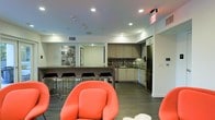 This single-floor space combines a fully equipped kitchen, a bar-style counter, and a comfortable lounge area in one open-plan layout. The kitchen is outfitted with dark stainless steel appliances, including a large refrigerator and double wall ovens, complemented by taupe cabinetry and a decorative mosaic tile backsplash that adds texture and visual interest. Ample upper and lower cabinets provide generous storage, while the clean countertops maintain a streamlined aesthetic.

A dark-topped bar counter separates the kitchen from the lounge area, flanked by a row of sleek black metal barstools that offer casual seating for socializing or informal dining. Recessed lighting throughout the ceiling provides consistent, even illumination across the entire space.

The lounge area is distinguished by bold, curved red chairs that create a vibrant contrast against the neutral walls and dark hardwood-style flooring. The overall design balances modern functionality with energetic pops of color, resulting in a welcoming and stylish communal environment well-suited for gathering, dining, or relaxing.