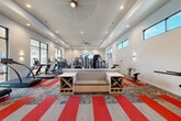 This spacious fitness center offers a comprehensive workout environment designed to accommodate a wide variety of exercise routines. The room features an elegant tray ceiling with recessed lighting and a large ceiling fan, creating a bright, comfortable atmosphere. Bold red and gray patterned carpet tiles add a dynamic visual element to the floor, energizing the space throughout.

The cardio zone is well-appointed with multiple treadmills, elliptical machines, stair climbers, and stationary bikes, all arranged along the perimeter to maximize open floor space. Floor-to-ceiling glass panel walls and large black-framed windows flood the interior with natural light and provide views of the outdoor pool area and surrounding grounds. A central seating bench with built-in cubby shelving serves as a functional divider between workout zones.

The strength training area includes a full rack of dumbbells, adjustable benches, medicine balls, and a multi-station cable and weight machine system. A dedicated rowing machine and additional specialty equipment further diversify the training options. Wall-mounted flat-screen televisions are positioned throughout the space for entertainment during workouts. Wall sconces complement the recessed lighting, and a mosaic tile accent partition adds a modern decorative touch. The overall design is clean, modern, and purpose-built for fitness.