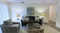 This single-floor space centers around a thoughtfully arranged living area that blends contemporary and coastal-inspired design elements. The room features a dark upholstered sofa accented with light blue and white throw pillows, creating a pleasing contrast against the soft, neutral-toned walls. Flanking the sofa are two glass-top side tables, each topped with round-based ceramic lamps that cast a warm, ambient glow throughout the space. A woven wicker coffee table serves as a focal point in front of the sofa, styled with decorative wooden bottle vases and a rustic tray, adding layers of texture and visual interest. A patterned accent chair or ottoman is visible to the left, contributing additional seating and a touch of bold pattern to the otherwise understated palette. Abstract wall art in muted blues and grays hangs above the sofa, reinforcing the room's cohesive, contemporary aesthetic. The flooring appears to be light-toned carpet, adding warmth underfoot. The overall ambiance is relaxed yet curated, with careful attention to symmetry, texture, and tonal harmony throughout the living space.
