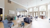 This single-floor community clubhouse is designed with an open, airy layout that immediately impresses with its soaring ceilings and an abundance of natural light. The main lounge area is anchored by a striking tray ceiling with a bold teal accent, crown molding, and light hardwood flooring that flows seamlessly throughout the space. Generous seating arrangements—featuring navy leather chairs, patterned accent chairs, and coordinated ottomans—are thoughtfully arranged across multiple distinct zones, creating both intimate gathering spots and larger social areas. Tall arched windows and sets of French doors line the walls, offering expansive views of the outdoor pool and lush landscaping, while inviting natural sunlight deep into the interior. A dedicated lounge nook equipped with wall-mounted televisions and comfortable seating provides a relaxed setting for casual viewing. The space also includes a well-appointed kitchenette featuring a mosaic penny-tile backsplash, a single-basin sink, a Keurig coffee station, and under-counter refrigeration. A dining area with a high-top table positioned in front of floor-to-ceiling gridded windows frames picturesque poolside views. Throughout, the warm neutral palette, decorative wall art in teal and gold tones, and refined architectural details combine to create a welcoming and sophisticated communal environment.