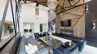 This sophisticated single-floor space makes an immediate impression with its soaring double-height ceilings and bold contemporary design. The centerpiece of the main lounge is a cascading cluster of oversized spherical chandeliers, crafting a dazzling focal point that draws the eye upward to a geometric, wood-paneled ceiling with dark metal accents — a striking architectural detail that adds warmth and texture to the expansive volume above.

Anchoring one wall is a dramatic floor-to-ceiling feature wall clad in richly toned wood panels framed by angular black metal inlays, housing a wide-format wall-mounted television and a sleek, linear gas fireplace below. This combination creates an inviting yet modern gathering point within the room.

The lounge area is furnished with plush gray sectional sofas and deep navy blue accent chairs arranged around a minimalist metal coffee table, all set atop a patterned area rug. A large abstract painting adds an artistic touch, complementing the overall contemporary aesthetic.

The open layout connects seamlessly to adjacent areas, including what appears to be a dining or co-working space visible in the background, enhancing the sense of flow and versatility throughout the space. Natural light filters in through high windows, amplifying the bright, airy atmosphere.