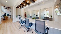 Located at 13695 Lead Ln in Lady Lake, Florida, this contemporary single-floor office space is thoughtfully designed to accommodate a variety of work styles. The open layout centers around a striking marble-topped conference table surrounded by modern ergonomic mesh chairs, making it ideal for collaborative meetings or focused group work. A wall-mounted display screen enhances the space's functionality for presentations.

Overhead, a standout architectural feature draws the eye: exposed dark wood ceiling beams are adorned with star-shaped pendant lights, adding warmth and elegance to the otherwise clean, white aesthetic. Light hardwood flooring throughout the main area contributes to the bright, airy atmosphere, complemented by abundant natural light streaming through large windows that offer views of palm trees and the surrounding landscape.

Along one wall, a continuous white countertop workstation provides additional seating for individual productivity, paired with open shelving and decorative accents. Private office pods enclosed with frosted glass sliding doors line the perimeter, offering quieter, more focused work environments, each equipped with a monitor, built-in desk, and its own natural light source. Patterned carpet tiles within the pods contrast beautifully with the main area's hardwood floors, creating clear spatial distinction throughout this well-appointed workspace.