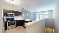 This single-floor unit showcases a contemporary open-concept layout that seamlessly connects the kitchen and living areas. The kitchen is a standout feature, equipped with dark espresso cabinetry, a white marble-style quartz peninsula with bar seating, and a full suite of stainless steel appliances including a refrigerator, range, microwave, and dishwasher. A white subway tile backsplash and pendant lighting above the peninsula add refined detail. The peninsula also includes a stainless steel sink, making it a highly functional workspace.

The living area extends naturally from the kitchen, finished with light wood-style flooring that runs throughout the unit. Large, multi-panel windows flood the space with natural light and are fitted with horizontal blinds for privacy control. A ceiling fan provides additional comfort and airflow.

The bathroom is cleanly designed with dark vanity cabinetry, a light countertop, a framed mirror, and a tiled shower-tub combination with chrome fixtures. The wood-style flooring continues here, maintaining visual consistency throughout the unit.

Overall, the space presents a clean, modern aesthetic with a neutral color palette of grays, whites, and warm wood tones, creating a cohesive and functional living environment.