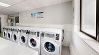 Located at 120 Torresdale Ave in Toronto, this shared laundry room is a well-organized and thoughtfully designed facility serving building residents. The space is bright and clean, featuring a suspended tile ceiling with recessed skylight panels that provide ample overhead lighting throughout. Light-toned large-format floor tiles contribute to the room's fresh, modern aesthetic.

The laundry area is generously equipped with multiple front-loading washing machines and top-loading dryers arranged in rows, offering considerable capacity for residents. A centrally located digital payment kiosk, mounted on a warm wood-panelled column, provides convenient card-based payment. Instructional signage is posted throughout to guide users on machine operation and laundry best practices.

A built-in shelving unit along one wall houses clearly labelled recycling and waste bins, supporting organized disposal. A textured accent wall adds a subtle design element to the space. Natural light filters in through a window on one side, further brightening the environment.

A dedicated waiting area features a row of teal upholstered chairs set against a clean white wall, offering a comfortable spot for residents while their laundry cycles run. The room is accessed through a secure grey door, ensuring controlled entry. Overall, this laundry facility is clean, functional, and well-maintained, providing residents with a practical and comfortable shared amenity.
