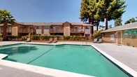 Located at 90 Lund Ave in Hayward, California, this outdoor communal pool area serves as a central gathering space within a residential complex. The centerpiece is a generously sized swimming pool with clear turquoise water, set within a wide, open concrete deck that provides ample room for lounge seating and relaxation. Surrounding the pool area is a well-maintained landscape featuring mature evergreen and palm trees that lend shade and a sense of privacy, along with neatly trimmed hedgerows and seasonal flowering plants that add color and vibrancy to the setting. The multi-building residential complex frames the pool on multiple sides, with units featuring private balconies and wood-accented exterior facades that contribute to a warm, suburban aesthetic. The architecture reflects a classic California residential style, with sloped rooflines, neutral earth-tone exteriors, and wood detailing. Under the abundant California sunshine, the pool area is an inviting outdoor retreat, offering residents a resort-like environment steps from their homes. The combination of lush greenery, open sky, and well-kept surroundings creates a relaxed, comfortable ambiance throughout the space.