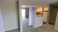 This single-floor residential unit offers a practical and well-organized layout designed to maximize both comfort and functionality. The living area is finished with wall-to-wall carpeting, creating a warm and inviting atmosphere, and benefits from natural light streaming in through a sliding glass door that opens to an outdoor balcony or patio area. A mirrored sliding closet door is prominently positioned along one wall, adding depth to the space while providing generous storage with a built-in hanging rod. Adjacent to the living area, the kitchen is accessible via an open pass-through counter with a granite-style surface, promoting a connected and social feel between the two spaces. The kitchen is well-appointed with white cabinetry, a white refrigerator, an electric range with overhead ventilation, and dark flooring that contrasts neatly with the lighter tones throughout. The neutral wall colors and clean finishes give the entire unit a bright, move-in-ready appearance. The thoughtful open-concept design between the kitchen and living spaces, combined with the natural light and practical storage solutions, makes this a comfortable and efficient living environment.