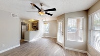 This single-story space offers a thoughtfully designed open-concept layout that connects the living area to the kitchen. The living room is finished with light wood-look hard flooring that runs throughout, contributing to a clean, cohesive aesthetic. A multi-blade ceiling fan with an integrated light fixture is centrally mounted on the textured ceiling, providing both airflow and ambient lighting. Multiple windows fitted with horizontal blinds allow natural light to filter into the living space, creating a bright and airy atmosphere.

The kitchen is partially open to the living area via a raised breakfast bar counter, offering a functional pass-through design. Dark cabinetry lines the kitchen walls, complemented by stainless steel appliances including a refrigerator and microwave. A decorative tile backsplash adds a visual accent to the cooking area.

A covered patio extends the living space outdoors, supported by textured stucco columns and bordered by a low wall. The patio overlooks a parking area shaded by mature trees, offering a natural backdrop. The exterior construction features textured stucco finishing consistent with the regional architectural style.