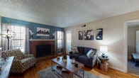 This single-story residence offers a well-organized layout that connects several distinct living areas. The living room serves as the focal point of the home, anchored by a full brick fireplace with a classic mantel and a bold steel-blue accent wall that adds visual depth. Hardwood-style flooring runs throughout the main areas, and a glass-paned door provides access to an outdoor space while allowing natural light to filter in. A textured ceiling is consistent throughout the unit.

The kitchen is efficiently designed, featuring warm oak cabinetry providing generous storage, dark granite-style countertops, stainless steel appliances including a range and dishwasher, and a black refrigerator. A breakfast bar separates the kitchen from an adjacent dining nook, which is brightened by a window and accommodates a bistro-style table for intimate dining.

The bedroom offers a carpeted floor, a window with blinds, and neutral wall tones that create a restful atmosphere. The full bathroom includes a single-sink vanity with dark countertop, wood cabinetry, hardwood-style flooring, and a shower/tub combination with a curtain enclosure.