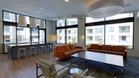 This single-floor space showcases a sleek, contemporary design centered around an expansive open-plan lounge area. Large, floor-to-ceiling windows along multiple walls flood the interior with abundant natural light while offering views of the surrounding building complex, including glimpses of palm trees that hint at a warm, urban setting.

The main seating area is thoughtfully arranged with bold burnt-orange sofas and accent chairs that provide a striking contrast against the light wood-toned flooring and neutral white walls and ceilings. Acrylic-legged coffee tables add a modern, airy touch to the furnishing scheme. Recessed ceiling lighting is complemented by stylish drum pendant fixtures and slim floor lamps, creating a layered and well-lit ambiance throughout.

A built-in wall unit anchors one end of the lounge, featuring an inset flat-screen television, a linear fireplace beneath it, and open shelving for display. This area also includes a pair of accent chairs for more intimate seating. Adjacent to the main lounge, a counter-height bar area with stools offers a casual space for dining or socializing, seamlessly integrated into the overall layout. The polished hardwood floors tie all zones together, contributing to the space's clean, cohesive, and inviting atmosphere.