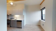 This single-floor space showcases a thoughtfully designed open-concept layout that seamlessly combines the kitchen and living areas into one cohesive environment. The kitchen is outfitted with sleek, flat-panel grey cabinetry, a white countertop, an undermount sink with a modern faucet, and a stainless steel dishwasher, all contributing to a clean, contemporary aesthetic. Warm wood beam accents at the ceiling add a touch of natural character, providing a subtle contrast to the otherwise crisp, white walls and ceiling. The light-toned wood-look flooring runs throughout the space, enhancing the sense of openness and warmth. A large black-framed window allows natural light to flood the living area, creating a bright and airy atmosphere. Under-cabinet lighting in the kitchen adds both functionality and ambiance. The open wall adjacent to the kitchen offers versatile space that can be adapted to a variety of living arrangements. A floor vent ensures efficient climate control throughout. The overall design is minimal and modern, making excellent use of the available space while maintaining a welcoming and functional feel.