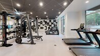 Located at 401 Oberlin Road in Raleigh, NC, this fitness center offers a generously sized, single-floor workout facility designed with both function and aesthetics in mind. The space is defined by a striking black drop-tile ceiling fitted with recessed lighting, crisp white walls, and light-toned flooring that together create a clean, modern atmosphere throughout.

A bold black-and-white geometric mural serves as a dramatic focal point, adding a contemporary artistic element to the interior. Full-length mirrors are strategically placed to enhance the sense of space and support proper form during training.

The facility is comprehensively equipped across multiple training zones. A central multi-function power rack anchors the strength training area, surrounded by a full dumbbell rack, a heavy boxing bag, battle ropes, TRX suspension trainers, adjustable benches, and cable machines. A dedicated cardio zone lines the perimeter, featuring treadmills, Peloton spin bikes, elliptical trainers, stair climbers, and a rowing machine.

Large floor-to-ceiling windows along one wall flood the space with natural light and provide open views of the surrounding Raleigh streetscape. A wall-mounted television is also present, supporting an engaging workout environment. The overall layout is spacious and well-organized, offering a professional-grade fitness experience.
