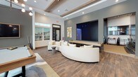 This single-story amenity space is thoughtfully designed to offer a diverse range of entertainment and social experiences. The main lounge area is anchored by soaring ceilings with exposed wood beam accents and recessed lighting, creating a warm yet contemporary atmosphere. Wood-look tile flooring runs throughout, adding texture and cohesion across the space.

The game lounge is well-appointed with a pool table, foosball table, and a chess table flanked by tan leather chairs, offering varied recreational options. A large wall-mounted flat-screen television with concrete drum stools provides casual viewing seating nearby. Multiple large windows allow natural light to filter in, brightening the neutral gray and white color palette.

A curved white sectional sofa faces an oversized display screen, creating a dedicated media viewing area ideal for group gatherings. Adjacent to the lounge, a sophisticated bar and kitchen area features a long dark countertop with bar seating, dark wood cabinetry, and a striking array of globe pendant lights overhead. Frosted glass double doors provide access to the kitchen.

A dedicated golf simulator room, finished in deep charcoal tones with a projection screen and textured carpet, rounds out the entertainment offerings, with an open pass-through window connecting it back to the main lounge.