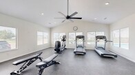 This dedicated exercise room is a comprehensively outfitted fitness space designed to support a full range of workout routines. The room features vaulted ceilings that lend an open, airy feel, complemented by recessed lighting and a large ceiling fan that ensures comfortable air circulation during workouts. Durable, speckled rubber flooring covers the entire space, providing both traction and impact absorption.

The cardio zone is well-stocked with multiple treadmills and elliptical trainers from Life Fitness, positioned along the windowed wall to take advantage of natural light and views of the adjacent outdoor pool area. A wall clock keeps exercisers on track during their sessions. On the opposite side of the room, a full multi-station cable and weight machine anchors the strength training area, accompanied by a set of adjustable benches and a tiered rack holding an organized selection of kettlebells and dumbbells. A wall-mounted mirror enhances the sense of space and allows for form-checking during exercises.

Large windows on multiple walls flood the interior with natural light and provide pleasant outdoor views, creating an inviting and motivating environment. The room is accessed via a standard interior door, and the overall clean, neutral design keeps the focus firmly on functionality and fitness.