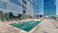 Located in the heart of Charlotte, NC, this rooftop amenity deck at 401 S. Graham Street offers a striking urban retreat set against the backdrop of the city's iconic skyline. The centerpiece of the space is a sparkling outdoor swimming pool, framed by a generous sun deck lined with teal lounge chairs — perfect for relaxing in the abundant natural sunlight. Adjacent to the pool, a bubbling hot tub provides a dedicated hydrotherapy area, adding to the resort-style atmosphere. The deck is enclosed by sleek glass railings that preserve unobstructed, panoramic views of the Charlotte skyline, including several of the city's most recognizable high-rise towers. Integrated planters with ornamental grasses add a touch of greenery and texture to the otherwise contemporary hardscape. The towering glass curtain wall of the building rises directly behind the pool area, reflecting the sky and city below and reinforcing the modern architectural character of the development. The open-air layout is thoughtfully designed to maximize both the sense of space and the visual connection to the surrounding urban environment, making this rooftop deck a standout amenity in downtown Charlotte.