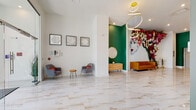 This single-floor leasing lobby is thoughtfully designed to make a striking first impression. The space is anchored by expansive marble-look porcelain tile flooring with warm gold veining that runs throughout, lending an air of sophistication and continuity across all areas.

The main reception area is bright and open, with crisp white walls providing a clean backdrop for carefully curated framed artwork, including a set of three white floral relief pieces. Plush gray accent chairs with vibrant orange cushions and a natural wood side table create an inviting waiting area with a contemporary flair.

A bold green accent wall defines a stylish alcove featuring a textured wooden credenza, a potted plant, and a gold-framed sunburst mirror — offering a refined focal point within the space. Adjacent to this, a large-scale face mural paired with a dramatic floral wall installation adds an artistic, vibrant energy to the room.

Toward the front of the lobby, floor-to-ceiling black-framed windows flood the space with natural light and provide clear views to the exterior. This area features a sleek linear fireplace set into the wall, a gold oversized wall clock, a cognac leather sofa, a mid-century orange accent chair, and a circular wood coffee table — all contributing to a warm yet modern aesthetic. A sculptural gold chandelier with interlocking rings completes the design overhead.