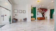 This single-floor leasing lobby is thoughtfully designed to make a striking first impression. The space is anchored by expansive marble-look porcelain tile flooring with warm gold veining that runs throughout, lending an air of sophistication and continuity across all areas.

The main reception area is bright and open, with crisp white walls providing a clean backdrop for carefully curated framed artwork, including a set of three white floral relief pieces. Plush gray accent chairs with vibrant orange cushions and a natural wood side table create an inviting waiting area with a contemporary flair.

A bold green accent wall defines a stylish alcove featuring a textured wooden credenza, a potted plant, and a gold-framed sunburst mirror — offering a refined focal point within the space. Adjacent to this, a large-scale face mural paired with a dramatic floral wall installation adds an artistic, vibrant energy to the room.

Toward the front of the lobby, floor-to-ceiling black-framed windows flood the space with natural light and provide clear views to the exterior. This area features a sleek linear fireplace set into the wall, a gold oversized wall clock, a cognac leather sofa, a mid-century orange accent chair, and a circular wood coffee table — all contributing to a warm yet modern aesthetic. A sculptural gold chandelier with interlocking rings completes the design overhead.