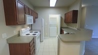 This single-floor space centers around a galley-style kitchen designed for practical, efficient use. The kitchen is lined on both sides with rich, dark wood cabinetry offering generous storage space above and below the countertops. Neutral-toned laminate counters run along both walls, providing ample workspace for meal preparation. White appliances are included, featuring an electric coil range with overhead microwave on one side and a full-size refrigerator on the other. A sink with countertop space is positioned along the far wall, complemented by a dishwasher tucked neatly beneath the counter. Light ceramic tile flooring runs throughout, contributing to a clean, low-maintenance aesthetic. A doorway at the end of the kitchen leads directly to a dedicated laundry alcove housing a stacked washer and dryer unit — a notable convenience. Overhead fluorescent lighting illuminates the kitchen corridor, keeping the space bright and functional. The neutral wall tones and practical layout make this a straightforward, well-equipped space suited for everyday living.