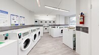 Located at 2050 Keele St in Toronto's Mount Dennis neighbourhood, this shared laundry facility is a compact yet efficiently organized space designed for high-volume use. The room features a modern, clean aesthetic with white walls accented by a horizontal band of dark grey tile, light wood-effect flooring, and bright fluorescent strip lighting that ensures excellent visibility throughout.

The facility is well-appointed with multiple front-loading washing machines arranged along the walls, complemented by a bank of stacked dryers, providing ample capacity for residents. Folding space is available atop the machines, offering convenience for users managing their laundry. A large window on one wall allows natural light to fill the space and provides views of the surrounding outdoor area, contributing to a pleasant environment.

Additional amenities include a payment kiosk for cashless transactions, informational signage posted on the walls for user guidance, and a utility sink. A security camera is mounted near the ceiling, ensuring the safety of the space. Access is provided through a secure door with a window panel. The layout is thoughtfully arranged to allow comfortable circulation, making this laundry facility a practical and well-maintained resource for the building's residents.