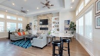 This single-floor space showcases a thoughtfully designed open-concept layout that seamlessly blends living and casual dining areas into one inviting environment. The focal point of the living area is a floor-to-ceiling stacked stone fireplace, flanked by symmetrical built-in shelving units with white cabinetry below — offering both display space and practical storage. A wall-mounted flat-screen television is integrated directly above the fireplace, creating a natural entertainment hub.

The tray ceiling adds architectural depth to the space, while recessed lighting and a ceiling fan ensure both ambiance and comfort throughout. Warm-toned hardwood flooring runs continuously across the space, grounding the bright, airy interior.

Adjacent to the living area, a curved granite-topped bar counter with bar stool seating provides a relaxed spot for casual dining or conversation. Large windows along the perimeter allow generous natural light to fill the room, enhancing the open and welcoming atmosphere.

Colorful artwork on the walls and vibrant accent pieces — including patterned area rugs and throw pillows — add personality and visual interest, balancing the neutral palette of the walls and furnishings. Lush indoor plants further enliven the space, contributing to its warm and inviting character.