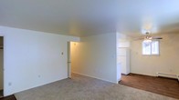 This single-floor space offers a clean, straightforward layout designed for functional living. The main living area is finished with wall-to-wall carpet in a neutral beige tone, providing a comfortable underfoot feel throughout. Smooth, light-colored walls and a plain ceiling contribute to a bright, open atmosphere, while multiple electrical outlets are conveniently distributed along the walls.

An open passage connects the living area to the kitchen, where the flooring transitions to a dark hardwood-style surface, creating a clear visual distinction between the two spaces. The kitchen is equipped with white cabinetry extending to the upper walls, offering ample storage, and a white refrigerator is visible, indicating a functional layout designed for practical use.

A doorway on the far side of the living area leads to an additional room, illuminated by a warm overhead light, suggesting a private space such as a bedroom or utility area. The overall aesthetic of the space is minimal and neutral, with white walls and simple finishes throughout, making it a versatile canvas well-suited for a variety of furnishing and décor styles.