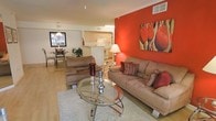 This single-floor space showcases a thoughtfully designed open-concept layout that effortlessly connects the living, dining, and kitchen areas into one cohesive and welcoming environment. The living room serves as the centerpiece, anchored by a striking red accent wall that adds warmth and personality to the space. Light hardwood flooring runs throughout, complementing the neutral tones of the furnishings and enhancing the bright, airy atmosphere. A glass-top coffee table and plush area rug anchor the seating arrangement, creating an inviting area for relaxation and conversation. The kitchen is accessible directly from the main living area and features light wood cabinetry, white appliances, and a convenient breakfast bar that doubles as a casual dining spot. Adjacent to the living room, a dedicated dining area is defined by a pendant light fixture overhead, offering an intimate setting for meals. Natural light filters through the windows, keeping the space feeling open and cheerful throughout the day. The consistent color palette — warm reds, soft tans, and light wood tones — lends the space a cohesive and polished look. Overall, this single-floor layout delivers a functional and stylish living experience with a bold, welcoming character.