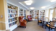This single-floor library is thoughtfully designed to provide a comfortable and functional space for reading and study. Built-in white shelving lines the walls from floor to ceiling, housing an extensive and well-organized collection of books across a variety of genres and subjects. The shelving units are neatly integrated into the architecture, giving the room a polished, classic appearance.

The seating arrangement balances both intimate and communal reading experiences. A pair of high-backed, cognac leather wingback chairs with contrast piping are positioned as a focal point, offering a refined and inviting spot for individual reading. Nearby, a dark wood dining-style table surrounded by upholstered side chairs provides space for group use or more structured reading sessions.

Overhead, flush-mounted drum pendant light fixtures provide consistent, warm illumination throughout the space. The neutral-toned carpet adds warmth underfoot while complementing the room's overall palette of cream, tan, and rich brown tones. Subtle decorative accents are interspersed among the books on the shelves, lending a curated, welcoming atmosphere to this well-equipped library.