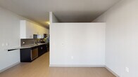 This single-floor residence offers a thoughtfully designed layout that balances open living with defined private spaces. Light wood-style tile flooring runs throughout the main areas, lending a clean, cohesive aesthetic. The entry hallway is lined with multiple doors, including a sliding barn-style door, and provides access to the various rooms within the unit.

The open-concept living and kitchen area is anchored by a well-appointed kitchen featuring dark lower cabinetry contrasted by crisp white upper cabinets, granite countertops, a mosaic tile backsplash, and stainless steel appliances including a refrigerator, microwave, dishwasher, and range. A breakfast bar extends from the counter, adding versatile functionality. The living area is generously proportioned, with light-filled windows along one wall providing outdoor views and natural light, complemented by a ceiling fan with integrated lighting and track lighting overhead.

The bedroom is comfortably sized to accommodate a queen bed and is finished with plush carpeting. The bathroom features a soaking tub with a full subway tile surround accented by a gray tile border, set atop wood-style tile flooring. The overall palette of neutral tones and modern finishes creates a clean, contemporary ambiance throughout.