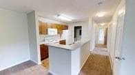 Located at 201 Eads Street in East Ridge, Tennessee, this single-floor residence offers a well-organized and functional layout designed for comfortable everyday living. The kitchen serves as a central highlight of the space, featuring warm honey-toned wood cabinetry with brushed hardware, white appliances including a range, over-the-range microwave, and refrigerator, and a peninsula with a neutral laminate countertop that provides additional prep and dining space. The kitchen floor is finished in wood-look flooring, adding warmth and easy maintenance underfoot. The peninsula opens directly toward the main living area, which is carpeted in a neutral beige tone that extends through the hallway and into the private rooms beyond. The hallway connects the living spaces to additional rooms, maintaining a clear and efficient flow throughout the home. Neutral gray-white walls and standard flush-mount ceiling lighting keep the interior feeling bright and clean. The overall ambiance is practical and move-in ready, with a straightforward design that allows residents to personalize the space with ease. The single-floor layout ensures accessibility and convenience throughout.