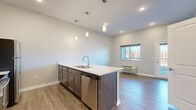 This well-designed single-floor unit offers a thoughtfully arranged layout that maximizes its compact footprint. The open-concept living and kitchen area serves as the heart of the space, anchored by a peninsula-style island with a light stone countertop, an undermount sink, and a stainless steel dishwasher. Three pendant lights hang above the island, providing focused task lighting. The kitchen is fully equipped with stainless steel appliances, including a refrigerator, electric range, and over-the-range microwave, complemented by dark wood cabinetry. Recessed lighting and neutral gray walls keep the area feeling bright and cohesive.

The living area features wood-look plank flooring that extends throughout the unit, contributing to a clean, modern aesthetic. A window provides natural light and an outdoor view, while a wall-mounted HVAC unit offers climate control. A dedicated laundry closet houses a stacked washer and dryer unit, adding everyday convenience.

The bathroom features a single-sink vanity with a light countertop, dark cabinetry, a toilet, and a bathtub. An adjacent closet includes built-in shelving and hanging rods, offering organized storage. The overall design is modern and functional, with a consistent neutral palette throughout.