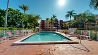 Located at 12375 SW 18th St in Miami, Florida, this outdoor pool area showcases a classic South Florida aesthetic defined by vibrant color, tropical greenery, and open-air leisure. The centerpiece is a generously sized rectangular swimming pool with crystal-clear turquoise water, framed by a terracotta tile deck that adds warmth and a Mediterranean character to the setting. Depth markers are visible along the pool's edges, indicating a gradual transition from shallow to deeper water. Surrounding the pool, a well-appointed deck offers ample space for relaxation, furnished with red lounge chairs and chaise lounges positioned to take full advantage of Miami's abundant sunshine. Shade options are provided by patio umbrellas and a dedicated poolside pavilion featuring a striking red exterior and a clay tile roof, which also serves as a pool house facility. The perimeter of the pool area is enclosed by a white fence and lush, mature tropical landscaping, including towering palm trees and dense canopy trees that provide a sense of privacy and a resort-like ambiance. The multi-story residential building, accented with bold red and neutral tones, rises in the background, complementing the colorful outdoor environment. This amenity space benefits from abundant natural light and open skies, characteristic of Miami's sunny climate.