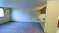 This single-floor space offers a straightforward, open layout designed for practical use. The main living area is covered in low-pile carpet, providing a neutral foundation that complements the light-toned walls throughout. A textured ceiling adds subtle visual interest overhead.

Along one side of the space, a built-in kitchenette is integrated into the layout, featuring a countertop with a stainless steel sink and wood-toned upper cabinetry. The counter space extends in an L-shaped configuration, offering a functional area for food preparation or utility tasks. The cabinetry provides modest storage above the work surface.

The open floor plan allows for flexible furniture arrangement within the main area, while the absence of interior partitions contributes to a sense of unobstructed space. Neutral wall colors and standard-height ceilings maintain a simple, clean aesthetic throughout. The space is well-suited for a variety of uses, with its minimalist design providing a blank canvas for customization. Electrical outlets are conveniently placed along the walls, supporting practical everyday use.