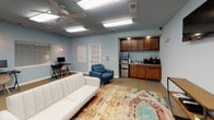 This single-floor space is thoughtfully designed to balance comfort and functionality, making it well-suited for a variety of professional or community uses. The interior is finished with soft blue-gray walls and a combination of carpet tile flooring in neutral and blue tones, creating a calm, welcoming atmosphere throughout.

The main open area serves as a versatile lounge and gathering space, anchored by a vibrant multicolored area rug that adds warmth and visual interest. Seating includes a white sofa, a blue upholstered armchair, and additional accent chairs, all arranged to encourage relaxed interaction. A wall-mounted flat-screen television and a half-moon console table are positioned along one wall, enhancing the space's media and lounge capabilities.

Along one side of the room, a built-in kitchenette features warm wood cabinetry, a countertop with a sink, a microwave, a beverage refrigerator, and a coffee station, providing convenient refreshment access without interrupting the flow of the space.

Toward the rear, a dedicated workstation area includes multiple computer desks with monitors and ergonomic seating, accommodating individual or small-group work needs. A traditional office desk with task chairs is also present, suggesting administrative or supervisory use.

Overhead, recessed fluorescent lighting and ceiling fans provide ample illumination and air circulation. Windows fitted with horizontal blinds allow natural light to filter in while maintaining privacy. An exterior door provides additional access to the space.