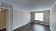This single-floor space presents a straightforward and functional interior with a neutral, move-in-ready aesthetic. The main living area features dark hardwood-style flooring that contrasts cleanly against the light grey walls, creating a balanced and understated look. White baseboards run along the perimeter of the room, adding a crisp finishing detail that complements the overall color palette.

The textured popcorn ceiling adds a classic touch, while crown molding along the upper edges of the walls provides a modest architectural detail that elevates the room's finish. A single window along one wall allows natural light to filter into the space, brightening the interior and offering a view of the surrounding exterior.

The room is open and unobstructed, allowing for flexible furniture arrangements to suit a variety of uses. Electrical outlets are conveniently positioned along the walls. The overall ambiance is clean, quiet, and neutral, making it a versatile canvas for personalization. The simple design and practical finishes give the space a functional character suited for everyday living.