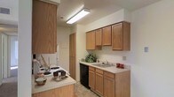 This single-floor space centers around a well-organized kitchen that combines functionality with a warm, approachable aesthetic. The kitchen features an abundance of medium-toned oak cabinetry, including both upper and lower cabinets that provide generous storage space throughout. A tall pantry-style cabinet offers additional storage, maximizing the use of vertical space. The countertops are finished in a light, neutral tone that complements the warm wood cabinetry, creating a cohesive and inviting atmosphere. A double-basin sink is positioned beneath the upper cabinets and is paired with a standard faucet, offering a practical workspace. The kitchen is equipped with a black dishwasher and an electric range cooktop, ensuring everyday convenience. Overhead, a large fluorescent flush-mount light fixture provides bright, even illumination throughout the space. The flooring features a neutral tile pattern that is both durable and easy to maintain. The white walls keep the space feeling open and clean, while the overall layout follows an efficient galley-style design that makes good use of the available space for cooking and meal preparation.