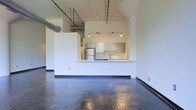 This single-floor space showcases an industrial loft aesthetic through a thoughtfully open layout. The most striking feature is the polished concrete flooring, which spans the entirety of the main living area, reflecting light and adding a sleek, urban character to the space. Soaring ceilings amplify the sense of openness, while exposed ductwork and structural elements overhead reinforce the industrial design language.

The kitchen is integrated into the open floor plan via a raised peninsula counter, which serves as a natural divider between the cooking and living areas without fully closing off either space. Upper cabinetry in a muted gray-green tone lines the kitchen walls, complemented by a subway tile backsplash that adds texture. Stainless steel appliances, including a full-size refrigerator, are visible within the kitchen area.

Track lighting mounted to the ceiling provides adjustable illumination throughout the space, offering both functional and atmospheric lighting options. The clean white walls throughout contrast with the rawer industrial elements, balancing the overall aesthetic and keeping the interior feeling bright and airy.