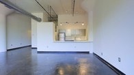 This single-floor space showcases an industrial loft aesthetic through a thoughtfully open layout. The most striking feature is the polished concrete flooring, which spans the entirety of the main living area, reflecting light and adding a sleek, urban character to the space. Soaring ceilings amplify the sense of openness, while exposed ductwork and structural elements overhead reinforce the industrial design language.

The kitchen is integrated into the open floor plan via a raised peninsula counter, which serves as a natural divider between the cooking and living areas without fully closing off either space. Upper cabinetry in a muted gray-green tone lines the kitchen walls, complemented by a subway tile backsplash that adds texture. Stainless steel appliances, including a full-size refrigerator, are visible within the kitchen area.

Track lighting mounted to the ceiling provides adjustable illumination throughout the space, offering both functional and atmospheric lighting options. The clean white walls throughout contrast with the rawer industrial elements, balancing the overall aesthetic and keeping the interior feeling bright and airy.