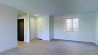 This single-floor space presents a contemporary, minimalist aesthetic defined by a neutral palette and clean architectural lines. The main living area is finished with light-toned hardwood flooring that extends throughout, contributing to a bright and cohesive feel underfoot. Smooth white walls and a flat ceiling with recessed lighting create a streamlined, modern environment that maximizes the sense of openness.

Natural light enters through a window fitted with horizontal blinds, providing both illumination and privacy control. The entryway area features a white interior door alongside a darker door, offering a subtle contrast that adds visual interest near the entry point of the space.

The overall layout appears open and unobstructed, lending itself well to flexible furniture arrangements. The absence of ornate detailing keeps the focus on the quality of the finishes, including the clean baseboards and smooth wall surfaces. The combination of recessed lighting and natural light ensures the space remains well-lit throughout the day. The overall ambiance is calm, clean, and move-in ready, suited to a variety of uses and interior styles.