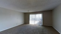 This single-floor space features a simply designed living area finished with wall-to-wall carpet in a neutral tone, contributing to a quiet and comfortable atmosphere. The room is characterized by its clean, understated aesthetic, with smooth beige walls complemented by white baseboards throughout. A textured popcorn ceiling adds a classic touch overhead, while a centrally placed ceiling vent ensures efficient climate control. The most prominent architectural feature is a large sliding glass door fitted with vertical blinds, which serves as the room's primary source of natural light. The door floods the interior with brightness and provides a direct visual connection to the outdoors, as well as convenient exterior access. The overall layout of the living area is open and unobstructed, allowing for flexible furniture arrangement. Electrical outlets are positioned along the walls at standard intervals, and a light switch is conveniently located near the entrance. The neutral color palette and soft carpeting create a calm, functional environment well-suited for everyday living.