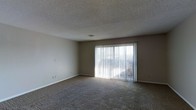 This single-floor space features a simply designed living area finished with wall-to-wall carpet in a neutral tone, contributing to a quiet and comfortable atmosphere. The room is characterized by its clean, understated aesthetic, with smooth beige walls complemented by white baseboards throughout. A textured popcorn ceiling adds a classic touch overhead, while a centrally placed ceiling vent ensures efficient climate control. The most prominent architectural feature is a large sliding glass door fitted with vertical blinds, which serves as the room's primary source of natural light. The door floods the interior with brightness and provides a direct visual connection to the outdoors, as well as convenient exterior access. The overall layout of the living area is open and unobstructed, allowing for flexible furniture arrangement. Electrical outlets are positioned along the walls at standard intervals, and a light switch is conveniently located near the entrance. The neutral color palette and soft carpeting create a calm, functional environment well-suited for everyday living.