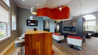 This vibrant, contemporary single-floor space is designed to impress, combining bold design choices with functional amenities throughout its open-plan layout. A dramatic, wave-shaped red ceiling feature anchors the central gathering area, drawing the eye upward while a modern ceiling fan adds a practical touch. Pendant lighting in textured grey tones complements the cool grey walls, creating a stylish contrast against the warm wood-toned flooring.

The lounge area is furnished with sleek white and red swivel chairs that echo the space's bold color palette, positioned around a linear stone-clad fireplace that serves as a striking focal point. A wall-mounted flat-screen television adds to the entertainment functionality of this zone. Nearby, a substantial warm-wood bar counter with dark metal bar stools provides a casual seating and gathering area, ideal for socializing.

An arched window stretches toward the high ceiling in the sitting area, flooding the space with natural light and offering views of lush greenery outside. Textured wall art, decorative pottery, and a black-and-white birch tree mural add curated artistic touches throughout.

The kitchen features dark espresso cabinetry, stainless steel appliances, mosaic tile backsplash, and light hardwood flooring. A separate seating nook with terracotta accent walls and woven-texture carpet rounds out this thoughtfully designed, multifunctional space.