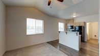 This single-floor space is defined by its vaulted ceiling, which lends an airy, open quality to the main living area. A ceiling fan with wooden blades is centrally mounted, providing both functional airflow and a subtle design accent against the angled ceiling. The living area features neutral-toned carpet that transitions into dark hardwood-style flooring, clearly delineating the boundary between the lounge and kitchen zones.

Large windows with horizontal blinds allow generous amounts of natural light to fill the space, contributing to a bright and welcoming atmosphere. The open-concept layout connects the living area directly to the kitchen, which is separated by a raised breakfast bar with corbel-supported shelving — a practical and visually appealing divider. The kitchen is equipped with black appliances, including a microwave and range, and is complemented by light wood cabinetry that provides ample storage. A secondary window in the kitchen area further enhances the natural lighting throughout. The overall color palette is clean and neutral, with white walls and light finishes creating a cohesive, understated aesthetic throughout the space.