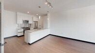 Located at 11036 Moorpark St in the North Hollywood neighborhood of Los Angeles, this single-floor unit showcases a clean, modern design with a cohesive aesthetic carried throughout every room. The open-concept living and kitchen area is bright and airy, with light hardwood-style flooring, crisp white walls, and large sliding glass doors that flood the space with natural light and provide access to an outdoor balcony with tree-lined views. The kitchen is outfitted with high-gloss white cabinetry, stainless steel appliances including a gas range, built-in microwave, refrigerator, and dishwasher, complemented by a peninsula with an integrated sink and light stone countertops — ideal for both cooking and casual dining. Pendant and track lighting add a refined touch overhead. The bedroom continues the modern theme with the same wood-style flooring, large sliding glass doors opening to a private balcony, and ample wall space. The hallway connects the living areas to the bedrooms and bathrooms with a smooth, logical flow. Bathrooms are finished with clean white surfaces, chrome fixtures, and a soaking tub-shower combination featuring elegantly tiled surround walls with a built-in niche. The overall ambiance is contemporary, minimal, and move-in ready.