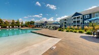 Located at 85 Washington St in Plainville, Massachusetts, this outdoor amenity space serves as a central gathering hub for its residential community. The centerpiece is a large, rectangular swimming pool with crystal-clear water, surrounded by an expansive stamped concrete sun deck outfitted with rows of lounge chairs and umbrella-shaded seating areas, ideal for relaxation and socializing during warmer months. A dedicated patio zone adjacent to the pool offers additional dining and lounge furniture beneath striped umbrellas, providing a comfortable outdoor living experience. The pool area is enclosed by a sleek black iron fence, ensuring both safety and a polished aesthetic. A single-story clubhouse or pool house building with large windows sits at the pool's edge, offering convenient access to amenities. Beyond the pool enclosure, the grounds open up to well-manicured green lawns interspersed with concrete walkways, ornamental plantings, and mature trees that provide shade and visual interest throughout the property. A dedicated playground area with slides and climbing structures is nestled within the landscaped grounds, catering to younger residents. The multi-story residential buildings visible in the background feature balconies, neutral siding, and stone accents, complementing the overall design of the community.
