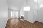 This two-story space is defined by its striking architectural height, most notably in the main living area where soaring ceilings and a dramatic volume of space create an open, airy atmosphere. A pendant light fixture hangs prominently, accentuating the vertical scale of the room. A carpeted staircase with a clean white railing leads to the upper level, while a tall window near the entry floods the transition space with natural light.

The living area features warm hardwood-style flooring, a white corner fireplace, and a bay window that invites abundant natural light and outdoor views. A back door with an adjacent window provides additional brightness and direct outdoor access.

The kitchen is efficiently designed with white cabinetry, a white countertop breakfast bar, recessed lighting, and stainless steel appliances including a refrigerator, range, and microwave. The layout opens toward the dining area, promoting a connected flow.

Upstairs, carpeted bedrooms offer a quieter, softer feel. One features vaulted ceilings and a closet, while another includes multiple windows with outdoor views. The bathroom is finished with a white vanity, white countertop, undermount sink with brushed nickel fixtures, a soaking tub, and hardwood-style flooring.