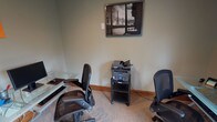 This business center is a thoughtfully appointed, single-floor workspace designed to meet a variety of professional needs. The entrance greets visitors with a classic dark-framed glass door flanked by sidelights, allowing natural light to flow in and offering a clear view of the landscaped grounds outside — setting a welcoming tone from the moment you arrive.

The main work area is equipped with glass-top desks, ergonomic mesh office chairs, a desktop computer with monitor, keyboard, and peripherals, and a multifunction printer on a dedicated rolling cart — providing all essential tools for productivity. The neutral, muted green walls and carpeted flooring contribute to a calm, focused atmosphere throughout.

A standout feature of the space is a cozy reading nook furnished with a cushioned armchair and a well-stocked bookshelf, positioned beside a beautiful stained glass window framed in warm wood. This area invites a moment of pause and reflection amid a busy workday. Decorative wall art, including framed black-and-white photographs with Parisian themes and a charming bicycle wall sculpture, add personality and character to the space. The warm wood baseboards and trim complement the overall aesthetic, lending a polished, professional feel to this practical and inviting business center.